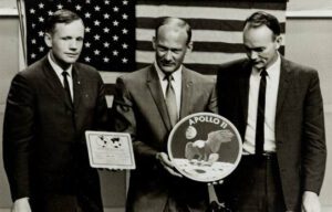 Posádka Apolla 11 (zleva Neil Armstrong, Edwin Aldrin a Michael Collins) poprvé ukazuje emblém mise letu; Neil Armstrong pak v ruce drží plaketu, která bude na lunárním modulu.